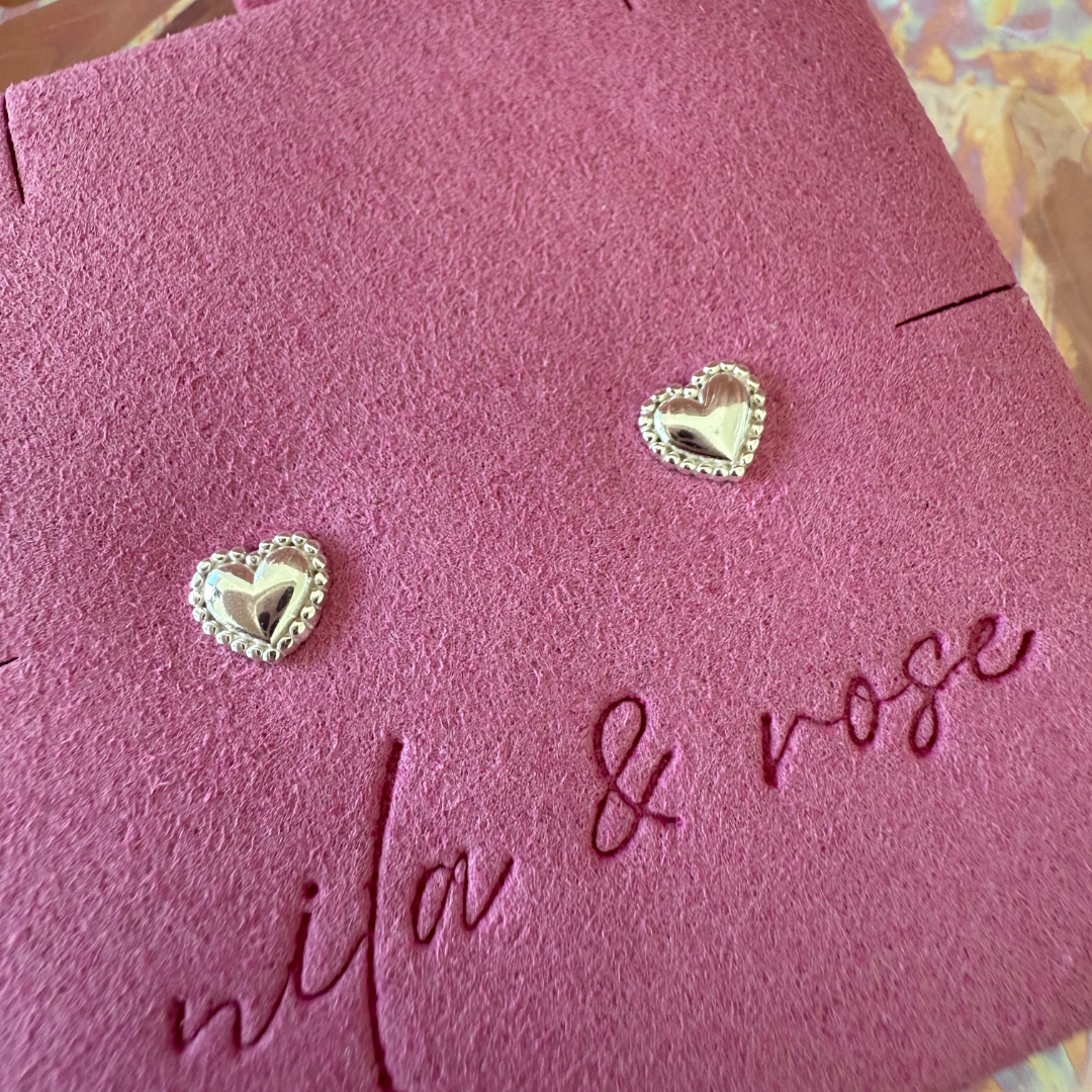 A pair of heart-shaped sterling silver stud earrings on a pink card with 'nila & rose' written in an elegant script.
