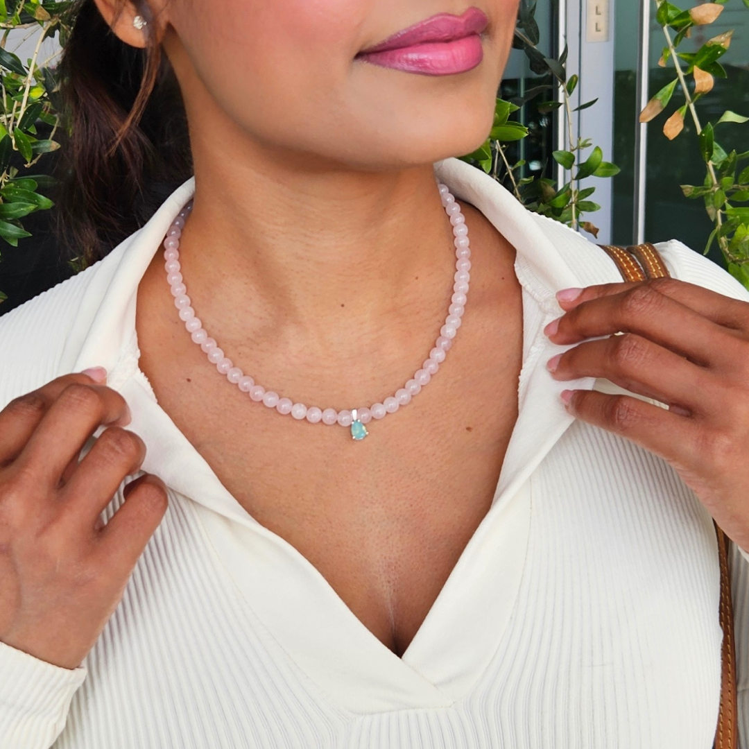 A person wearing a white v-neck sweater is showcasing a delicate necklace with soft pink rose quartz gemstones and a sea-green pendant.